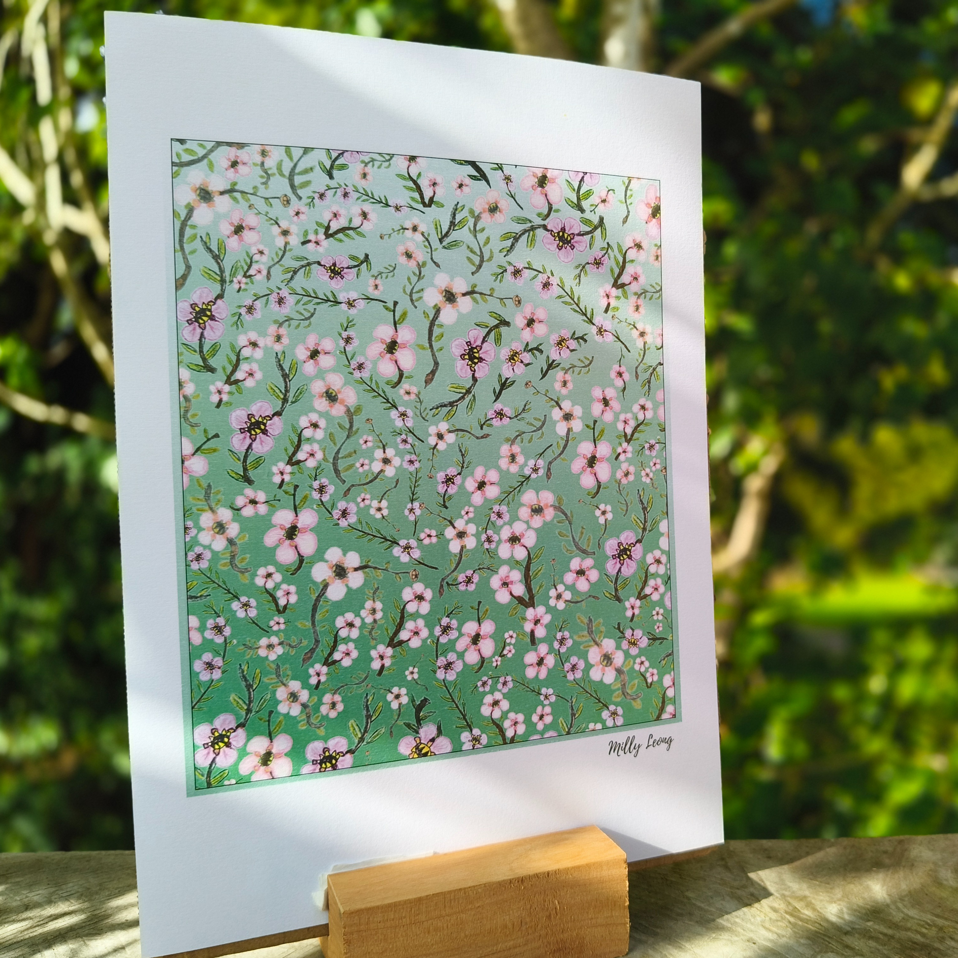 A4 art print with pink floral pattern on a green background, held by a wooden stand with a natural background.