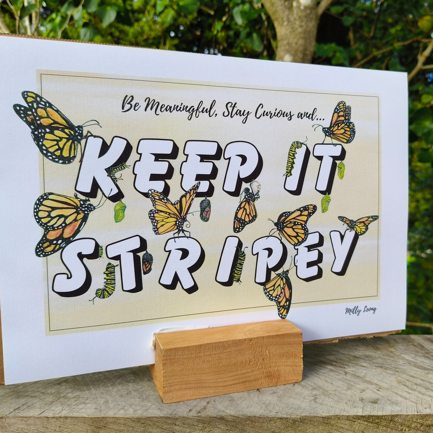 A4 art print with butterflies and text 'KEEP IT STRIPEY' on a wooden stand outdoors.